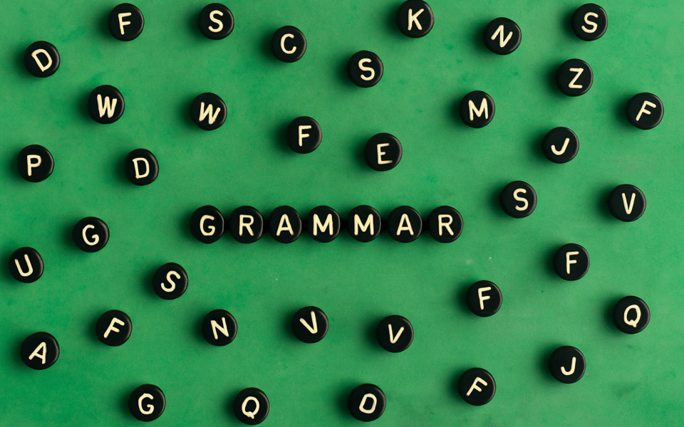 Czarne guziki z białymi literami ułożone na zielonym tle. W centrum widoczne słowo „GRAMMAR” ułożone z liter, otoczone przypadkowymi literami alfabetu rozrzuconymi wokół.